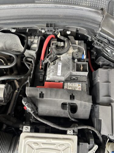 メルセデスベンツBクラス 補機バッテリー交換 修理時のエンジンルーム内バッテリー設置状態 メルセデス・ベンツBクラスのエンジンルーム内に搭載されている補機バッテリーの設置状態。カバーを外した状態で、配線や端子接続部分が確認できる作業前の様子。