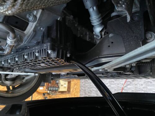 リフトアップしたメルセデスベンツEクラスの下回り。 オイルドレンから古いエンジンオイルを抜き取っている様子で、オイルパン周辺やアンダーカバーが確認できる作業中の写真。